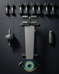 A flat-lay view of a black and gray gym weight bench setup on a dark speckled floor, surrounded by dumbbells, an ab roller, a water bottle, and resistance bands.
