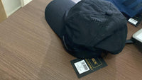 A black SoccerEx mesh face cap with a visible brand tag on a wooden surface.