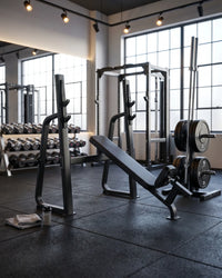 A black adjustable weight bench and weight stack rack set in a modern gym with large windows, dumbbells, and industrial-style ceiling lights.
