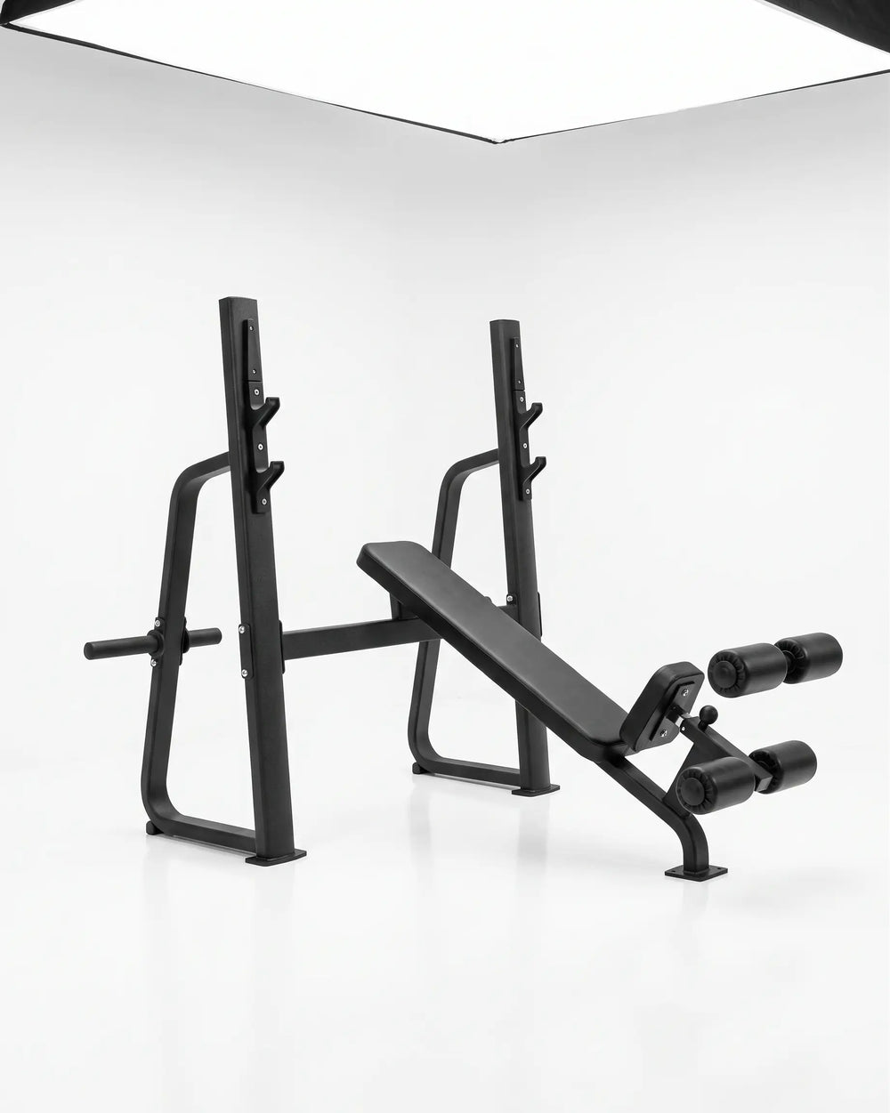 A black adjustable weight bench press with a safety guard and dual roller attachments, photographed in a clean white studio setting.