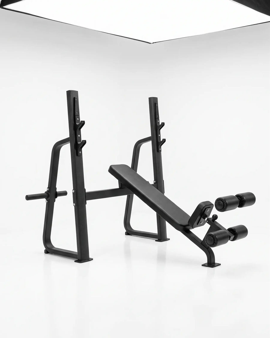 A black adjustable weight bench press with a safety guard and dual roller attachments, photographed in a clean white studio setting.