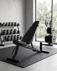 A black adjustable weight bench with a padded backrest and leg rest, positioned on a black gym mat inside a modern gym. Dumbbells are visible on a rack in the background, along with a barbell on a weight stack and a large window showing greenery outside.
