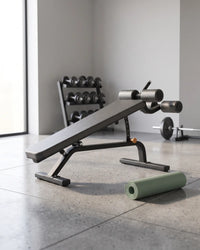 A black adjustable weight bench with a light green rolled yoga mat in the foreground, set in a minimalist gym with a dumbbell rack and barbell in the background.