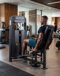 A man in a black t-shirt and teal shorts sits on a hip abductor gym machine inside a modern gym with wood paneling and other exercise equipment in the background.
